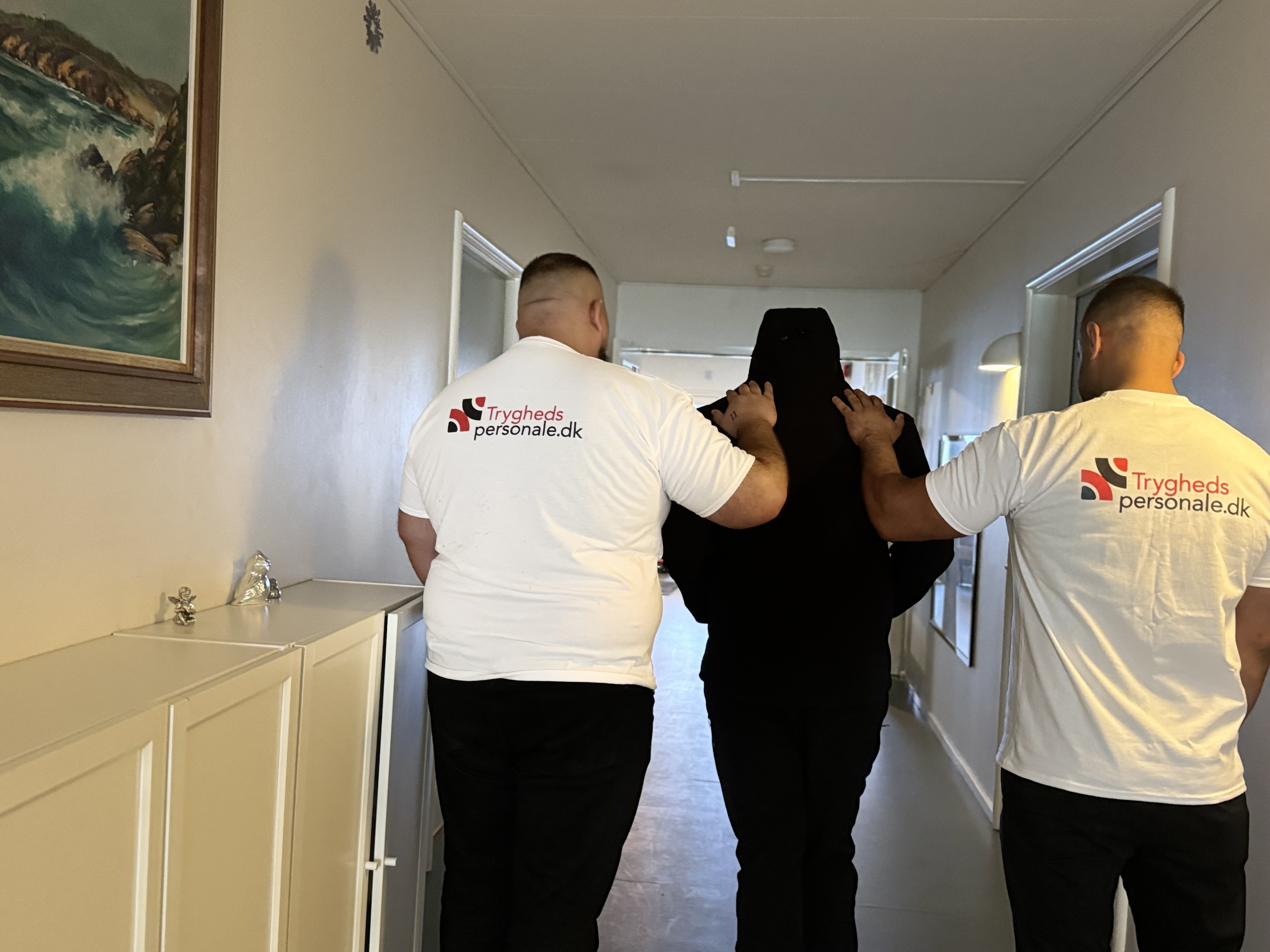 Tryghedspersonale medarbejdere eskorterer person i mørkt tøj